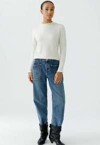 Lichtcrème trui met een gegolfde halslijn en lange mouwen, gecombineerd met losse blauwe jeans met grote zakken en ritsaccenten.