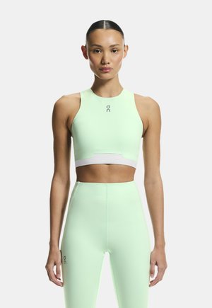 Žena oblečená v mátově zeleném sportovním krátkém topu a legínách s vysokým pasem, stojící čelem vpřed na světlém pozadí.