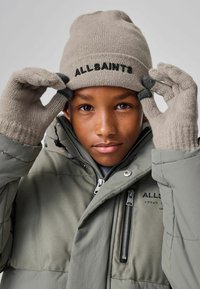 Beidzis adījuma beņķis ar "ALLSAINTS" logo, apvienots ar atbilstošām beidzis cimdiem. Modelis valkā pelēku pūkaino jaku ar rāvējslēdzēju detaļām.