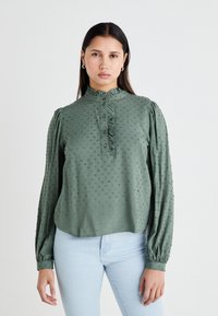 Blusa verde con colletto alto a volant, tessuto a trama con piccoli punti, chiusura a bottoni e maniche lunghe con polsini elasticizzati.