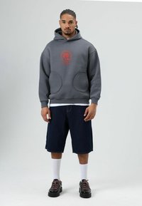 Grijze hoodie met kap en rode afbeelding, met grote voorzakken, gecombineerd met donkere denim shorts, witte sokken en zwarte schoenen.