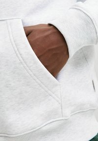 Gaišs pelēks sporta krekls ar ribotu aproci un kabatu; roka novietota kabatā, izceļot gludu ādas tekstūru.