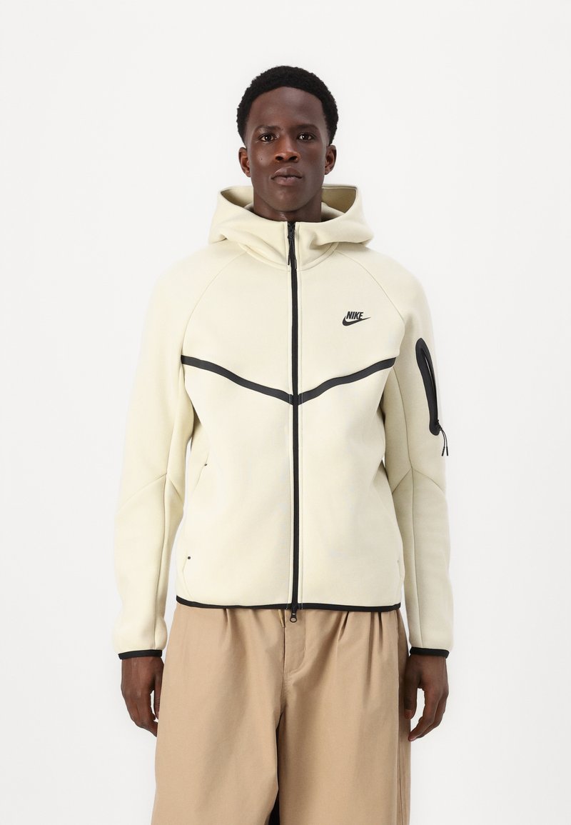 Veste à capuche zippée Nike en crème, avec des accents noirs, une poche latérale et une coupe ajustée avec une texture lisse.