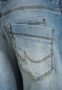 Jean en denim bleu clair avec une poche arrière dotée de coutures dorées contrastantes et d'une légère usure. Design simple et classique à cinq poches.