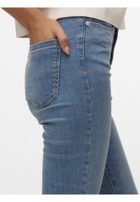 Gros plan d'une personne portant un jean en denim bleu clair avec la main droite dans la poche arrière, sur un fond uni.