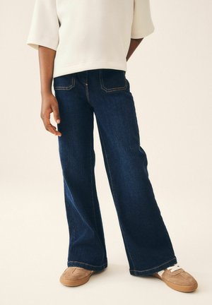 Person iført mørkeblå jeans med vide ben, beige sneakers med hvide snørebånd og en løs hvid top, stående mod en ensfarvet baggrund.