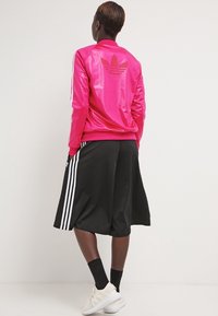 Rosa satinjacka med logga, svarta knälånga shorts med vita sidostripes och vita sportskor. Slät textur och sportig design.