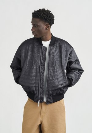 Ung mand med kort krøllet hår iført en sort quiltet bomberjakke over en hvid skjorte og beige bukser, hænderne i jakkelommerne, kigger mod venstre.