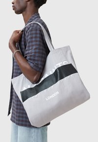 Borsa a spalla in canvas grigio con una fascia nera, caratterizzata dal testo "ALSAINTS" e "LONDON". Cinghia a spalla e texture liscia.