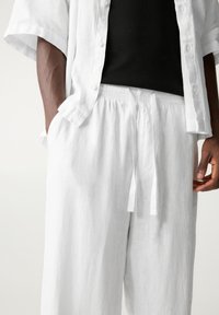 Homme portant un pantalon en lin blanc à cordon de serrage et une chemise assortie à manches courtes ouverte sur un débardeur noir côtelé, les mains détendues le long du corps.