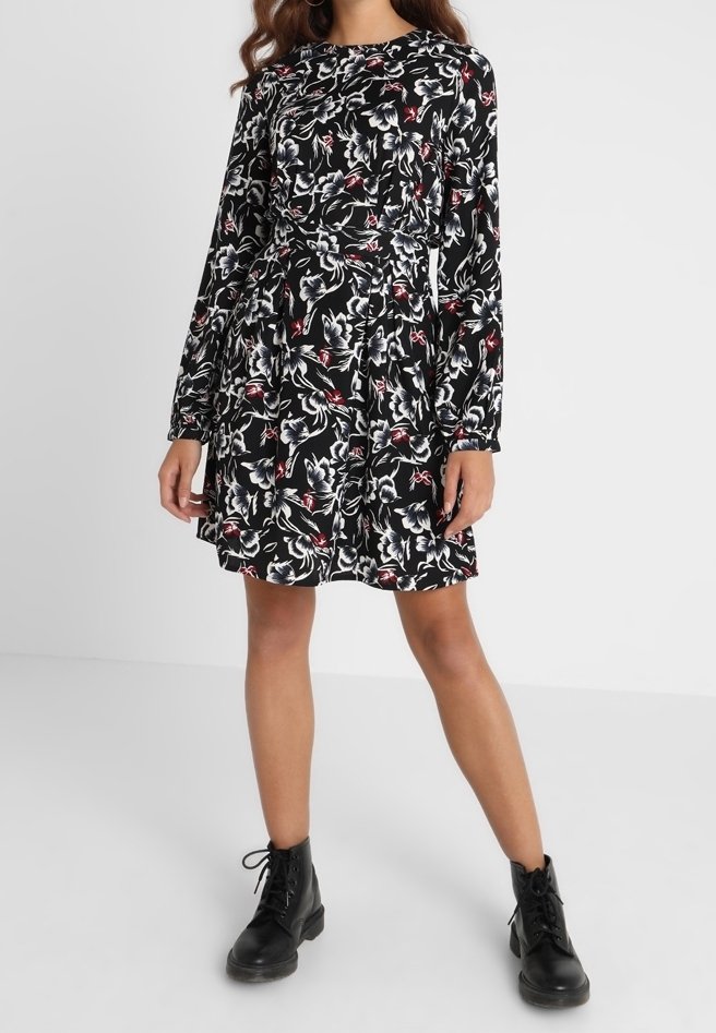 Robe noire à motifs floraux avec manches longues, dotée d'un corsage cintré et d'une jupe évasée. Le motif floral comprend des accents blancs et rouges. Portée avec des bottes noires.