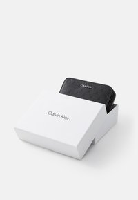Calvin Klein melnas ādas maciņš ar iegravētu rakstu, novietots baltā kastē ar minimālistisku fontu, kurā ir redzams zīmola nosaukums.