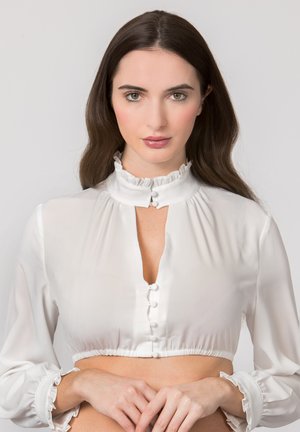 Junge Frau mit langen braunen Haaren, die eine weiße kurze Bluse mit Rüschenkragen und Manschetten trägt, steht vor einem einfarbigen Hintergrund.