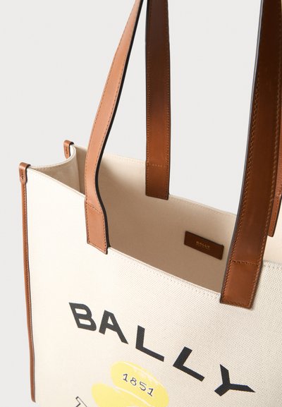 Τσάντα ώμου από καμβά κρεμ με καφέ δερμάτινες λαβές και λεπτομέρειες, με μαύρο κείμενο "BALLY" και κίτρινο γραφικό με "1851" στο μπροστινό μέρος.