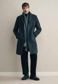 Abrigo de lana gris oscuro con solapas de muesca, combinado con una bufanda verde y pantalones negros. Zapatos negros completan el look. Diseño simple y moderno.