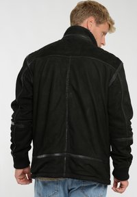 Sort ruskindjakke med ståkrave, struktureret stof og læderdetaljer ved skuldrene og ærmerne. Synlig syning.