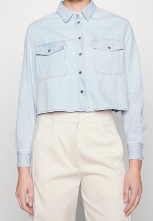 Chemise en denim bleu clair raccourcie avec deux poches sur la poitrine, portée avec un pantalon beige taille haute.
