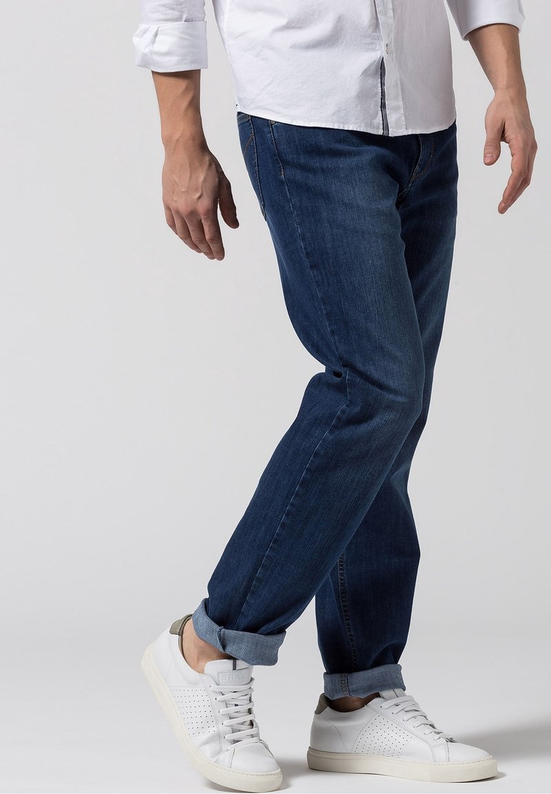 Jeans de mezclilla en azul oscuro, corte recto con puños enrollados. Combinados con zapatillas blancas que presentan detalles perforados y acentos grises.