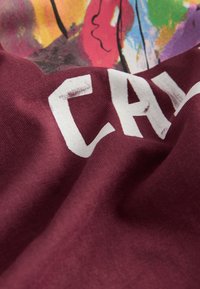 Tela de algodón marrón con una textura suave que presenta grandes letras blancas. El fondo muestra un diseño abstracto colorido.