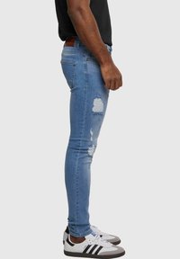 Heledad hall denim skinny teksad heledastusega, millel on sobiv kuju, viis taskut ja pruun nahkplekk vööl. Kaasa on pürgitud valgete tossudega.