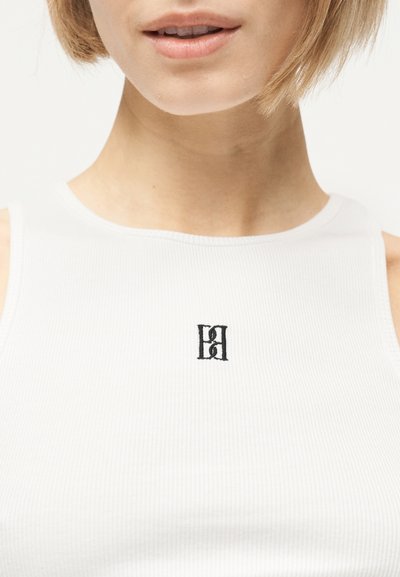 Κοντινό πλάνο γυναίκας που φοράει λευκό ριμπ κορμάκι με ένα μικρό μαύρο κεντημένο λογότυπο στο κέντρο του στήθους.