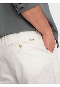 Pantalons en lin couleur beige clair avec une poche arrière à fermeture bouton et un petit logo métallique. Le tissu a une apparence texturée.