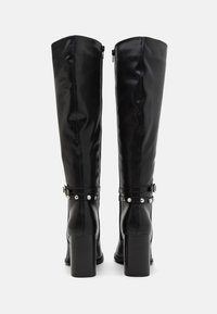 Botas negras de cuero suave hasta la rodilla, con tacón cuadrado, cierre de cremallera y cinturón decorativo con tachuelas plateadas.