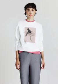 Felpa bianca con un design cropped, caratterizzata da una grafica rosa di una figura. Indossata sopra una maglietta rosa, abbinata a pantaloni grigi con texture.