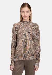 Kobieta w długim rękawie, z wysokim kołnierzem, nosząca bluzkę z bogatym brązowo-beżowo-zielonym wzorem paisley oraz gładkie brązowe spodnie.
