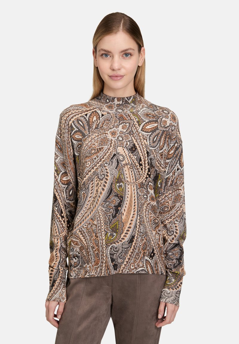 Kobieta w długim rękawie, z wysokim kołnierzem, nosząca bluzkę z bogatym brązowo-beżowo-zielonym wzorem paisley oraz gładkie brązowe spodnie.