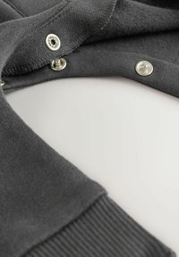 Grauer Sweatshirtstoff mit weicher Textur, ausgestattet mit Druckknöpfen aus silbernem Metall und einem gerippten Bündchen. Nahaufnahme des Kragenbereichs.