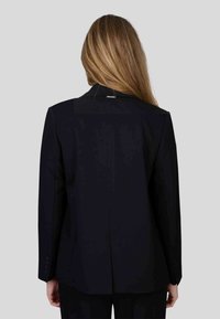 Blazer bleu marine ajusté avec une texture lisse, des revers nets, une fente arrière unique et des accents à quatre boutons sur les manches. Vue arrière simple.