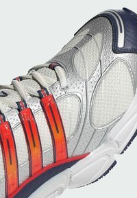 Silberner Sportschuh mit strukturierten Oberflächen, roten und orangefarbenen Akzenten und weißer Sohle. Verfügt über Schnürsenkel und Gummibesätze zur Unterstützung.