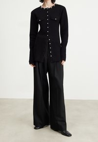 Persona vistiendo un largo cárdigan negro con botones de perla, pantalones anchos negros y zapatos de punta negros sobre un fondo claro.