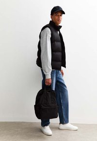 Czarna pikowana kamizelka na jasnoszarym swetrze, niebieskie dżinsy i białe trampki. Czarna plecak z przednią kieszenią, stojący pod gładką ścianą.