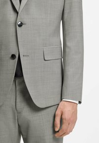 Veste et pantalon de costume gris clair avec deux boutons noirs, poche à rabat, manchette de chemise blanche visible et main détendue sur le côté.