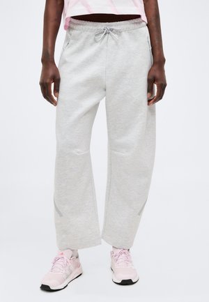 Persoon draagt lichtgrijze wijd uitlopende joggingbroek met trekkoord in de taille en roze sportschoenen, staand tegen een witte achtergrond.
