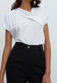 Top blanco de manga corta con escote drapeado y detalles fruncidos. Combinado con pantalones negros de cintura alta con un botón dorado. Textura suave.