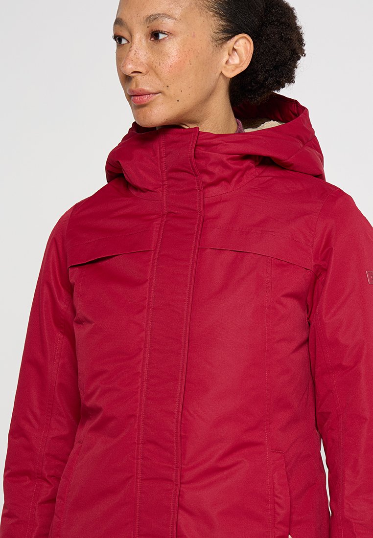 Frau mit lockigem Haar, die eine rote Winterjacke mit Kapuze trägt und seitlich vor einem schlichten Hintergrund schaut.