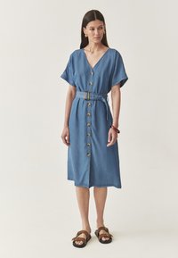 Denim midi-mekko, jossa on V-pääntie, lyhyet hihat, etupainon sulkeminen, vyötärölle sidottu vyö ja taskut. Pehmeä rakenne vaaleansinisessä värissä.