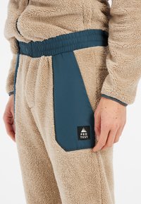 Prenda de forro polar beige con paneles de acento en azul verdoso. Presenta una superficie texturizada, cintura elástica y un parche de marca en el bolsillo lateral.