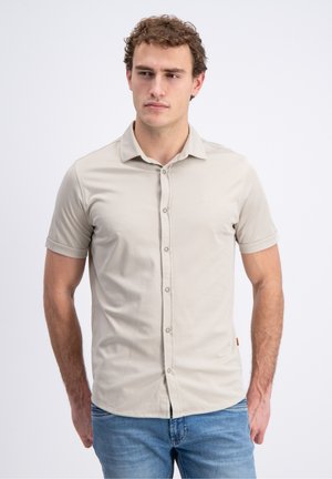 Mand med krøllet hår iført en beige kortærmet skjorte med knapper og blå jeans, stående med hænderne afslappet ned langs siderne.