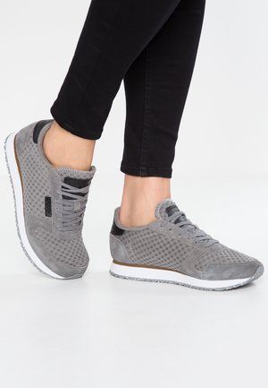 Persona con zapatillas de malla y ante gris con suelas blancas y detalles en negro, combinadas con pantalones ajustados negros, de pie sobre un suelo blanco.