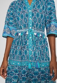 Abito blu con motivo floreale, maniche corte, dotato di una chiusura frontale a bottoni, vita con cordoncino e dettagli con pon-pon.
