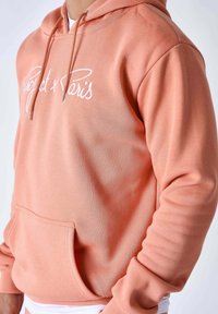 Sudadera color melocotón con capucha ajustable con cordón, bolsillo delantero y texto bordado "Project x Paris". Tejido suave y puños acanalados.