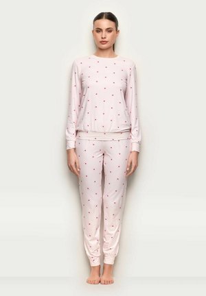 Donna in piedi a piedi nudi che indossa una maglia a maniche lunghe rosa chiaro e pantaloni con piccoli motivi a cuori rossi su uno sfondo semplice.