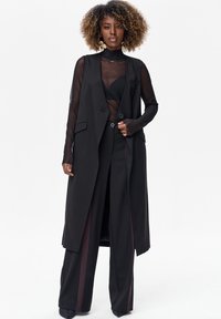 Vrouw met krullend haar draagt een doorschijnende zwarte top, een lange mouwloze zwarte vest, wijde zwarte broek met donkere zijstrepen, en grote oorbellen.