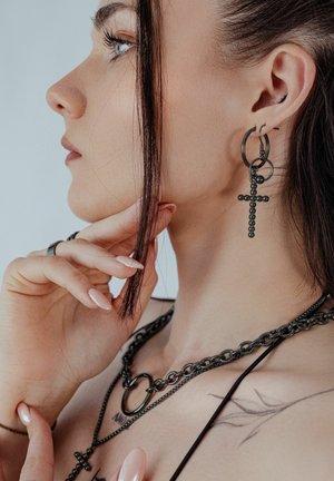 Junge Frau mit heller Haut, dunklen Haaren, trägt schwarze Kettenkette und schwarze Kreuzohrringe, berührt ihr Kinn mit gepflegter Hand.