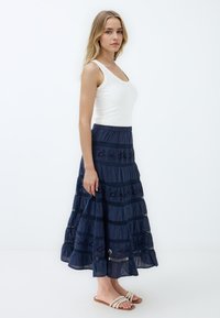 Jupe maxi bleu marine à volants, ornée de dentelle et à ourlet enroulé, associée à un débardeur blanc ajusté et des sandales beiges.