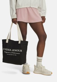 Borsa tote nera in canvas con testo bianco, manici crema, abbinata a una gonna sportiva rosa e sneakers chiare con dettagli arancioni.